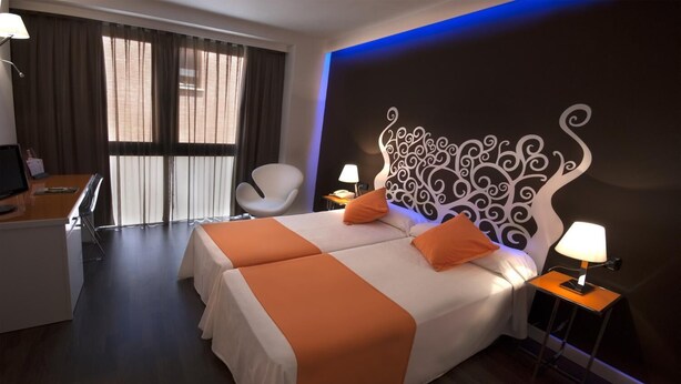Habitación de hotel con dos camas, un escritorio, una silla y un ventanal con cortinas.