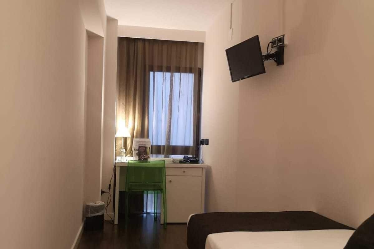 Una habitación de hotel con cama, escritorio y silla, televisión montada en la pared y ventana con cortinas.
