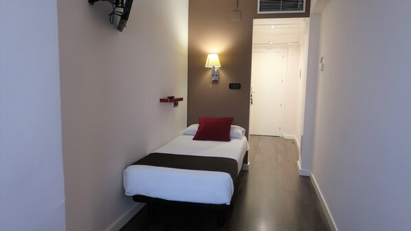 Un estrecho pasillo de hotel con una cama al final, una lámpara de pared y una puerta.