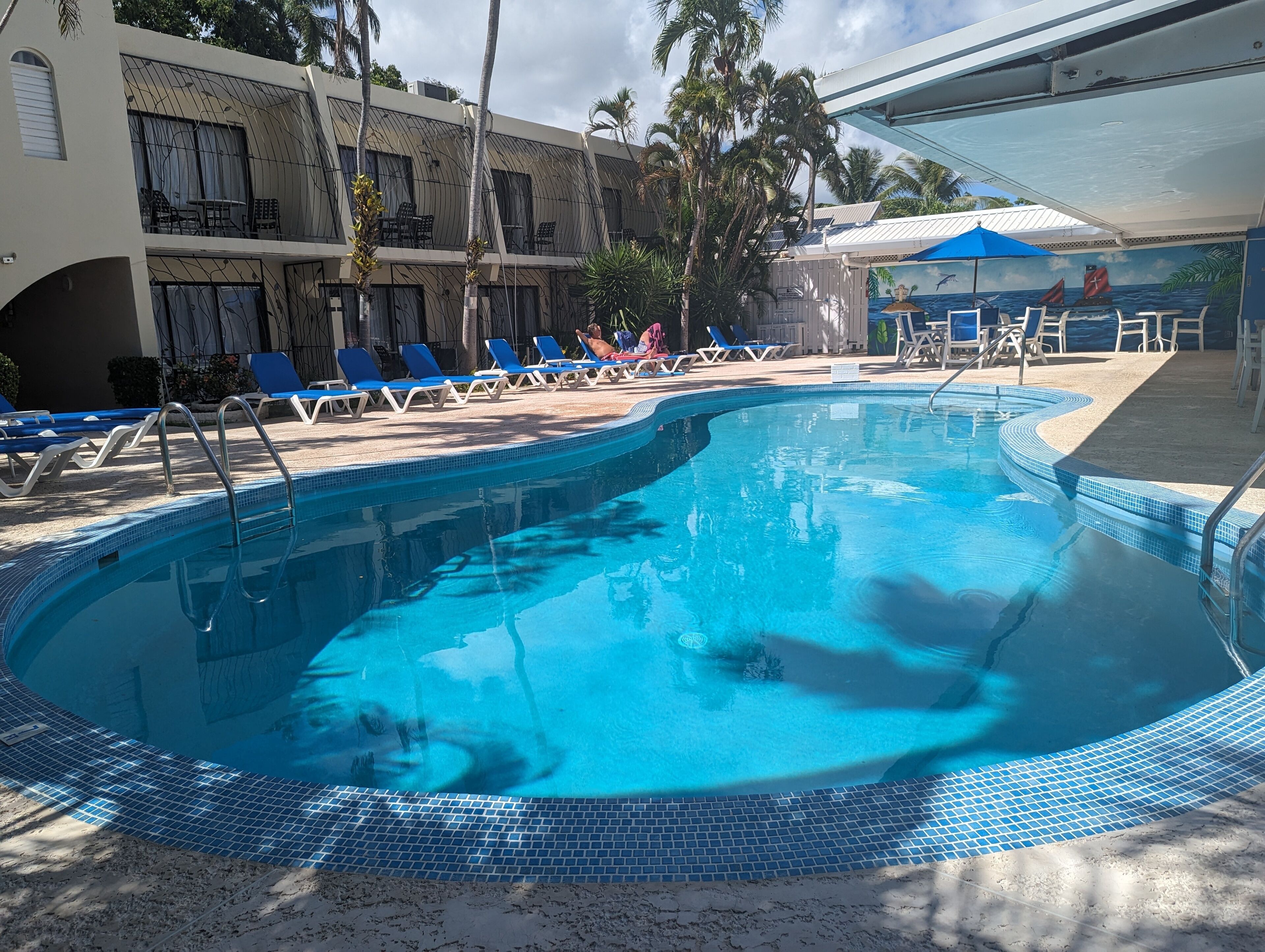 A swimming pool with blue-tiled edges, surrounded by blue lounge chairs and a building with balconies.
