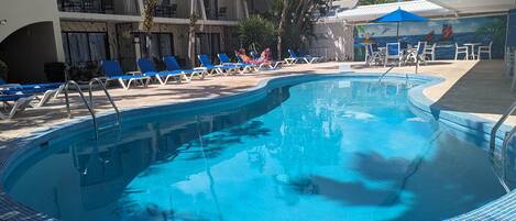 A swimming pool with blue-tiled edges, surrounded by blue lounge chairs and a building with balconies.