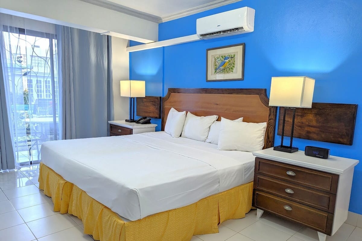 A hotel room with a large bed, two bedside tables, a nightstand, a wall-mounted air conditioner, and a window with curtains.