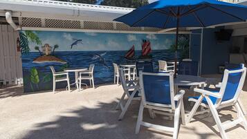 An outdoor seating area with white chairs and a blue umbrella, featuring a mural of a tropical scene with a boat and palm trees.