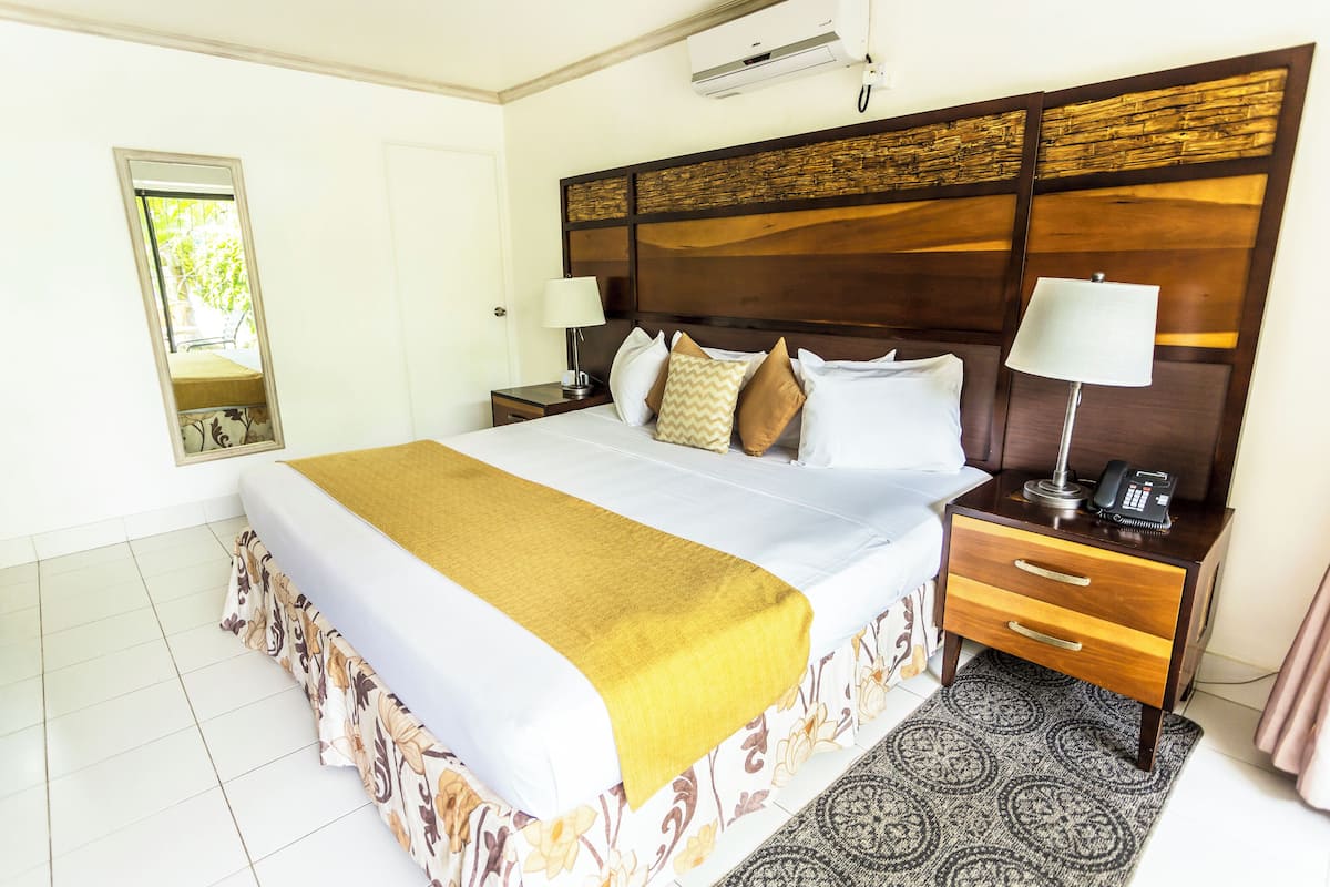 A hotel room with a large bed, a bedside table with a lamp, a mirror, and an air conditioning unit.