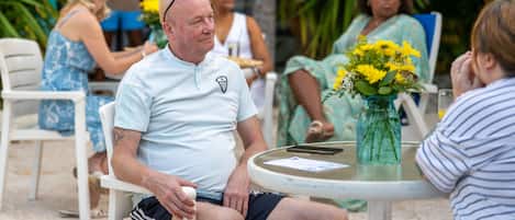 Un homme est assis à une table ronde à l’extérieur, avec un petit vase contenant des fleurs jaunes.