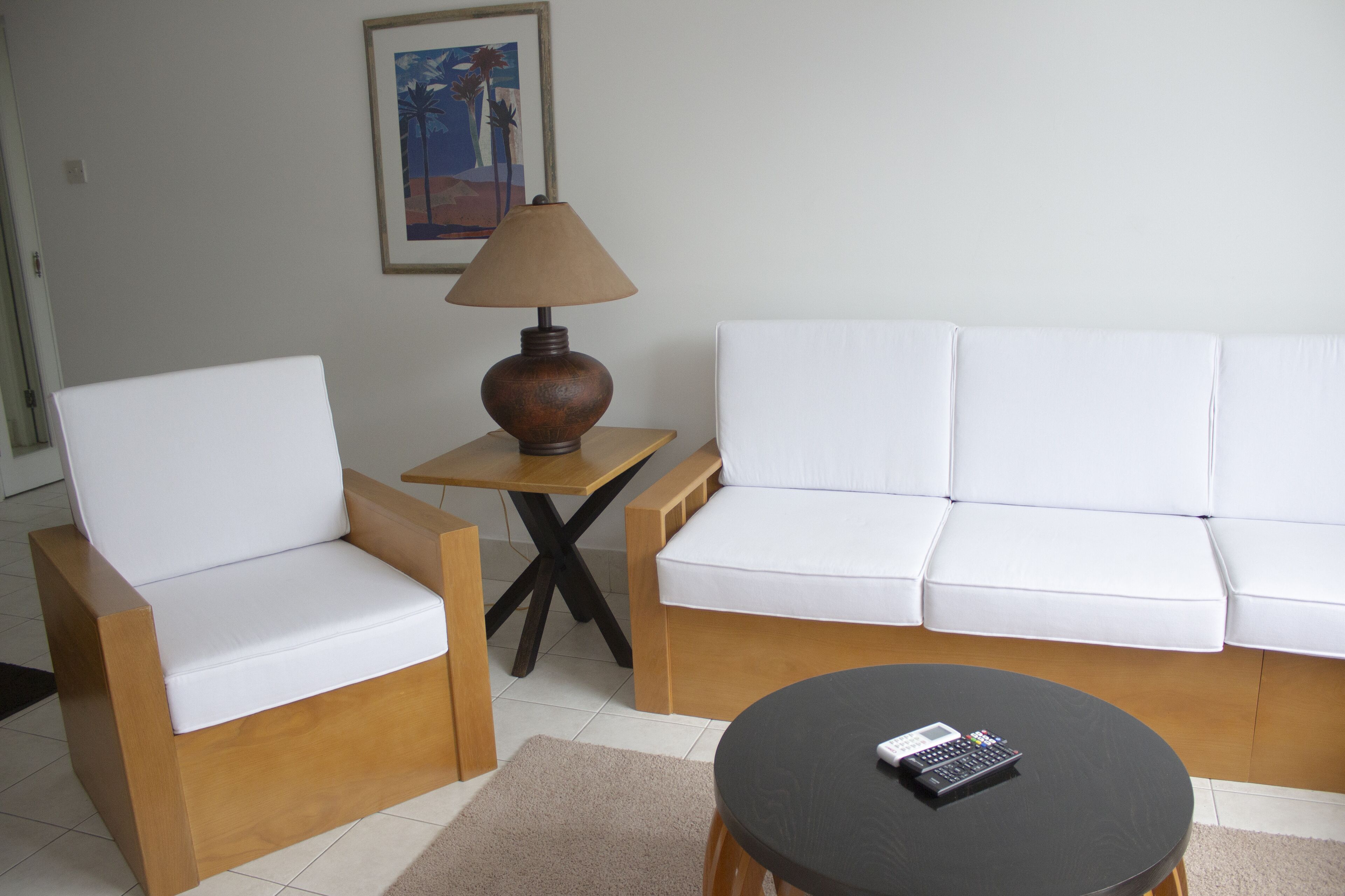A living room with a white sofa, two armchairs, a small wooden table with a lamp, and a coffee table with a remote control.
