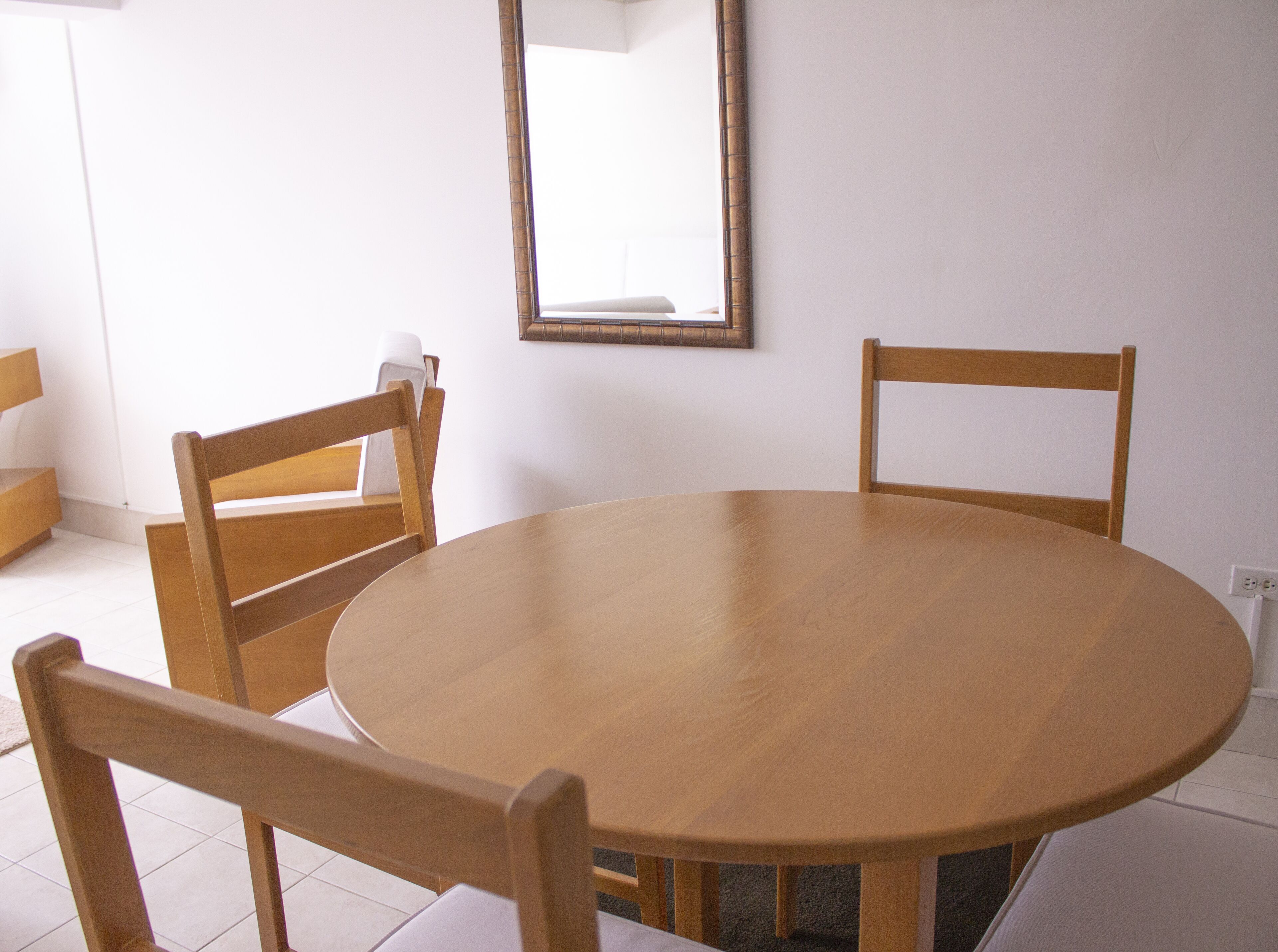 Una mesa redonda de madera con cuatro sillas de madera alrededor, una pared blanca y un espejo sobre la mesa.