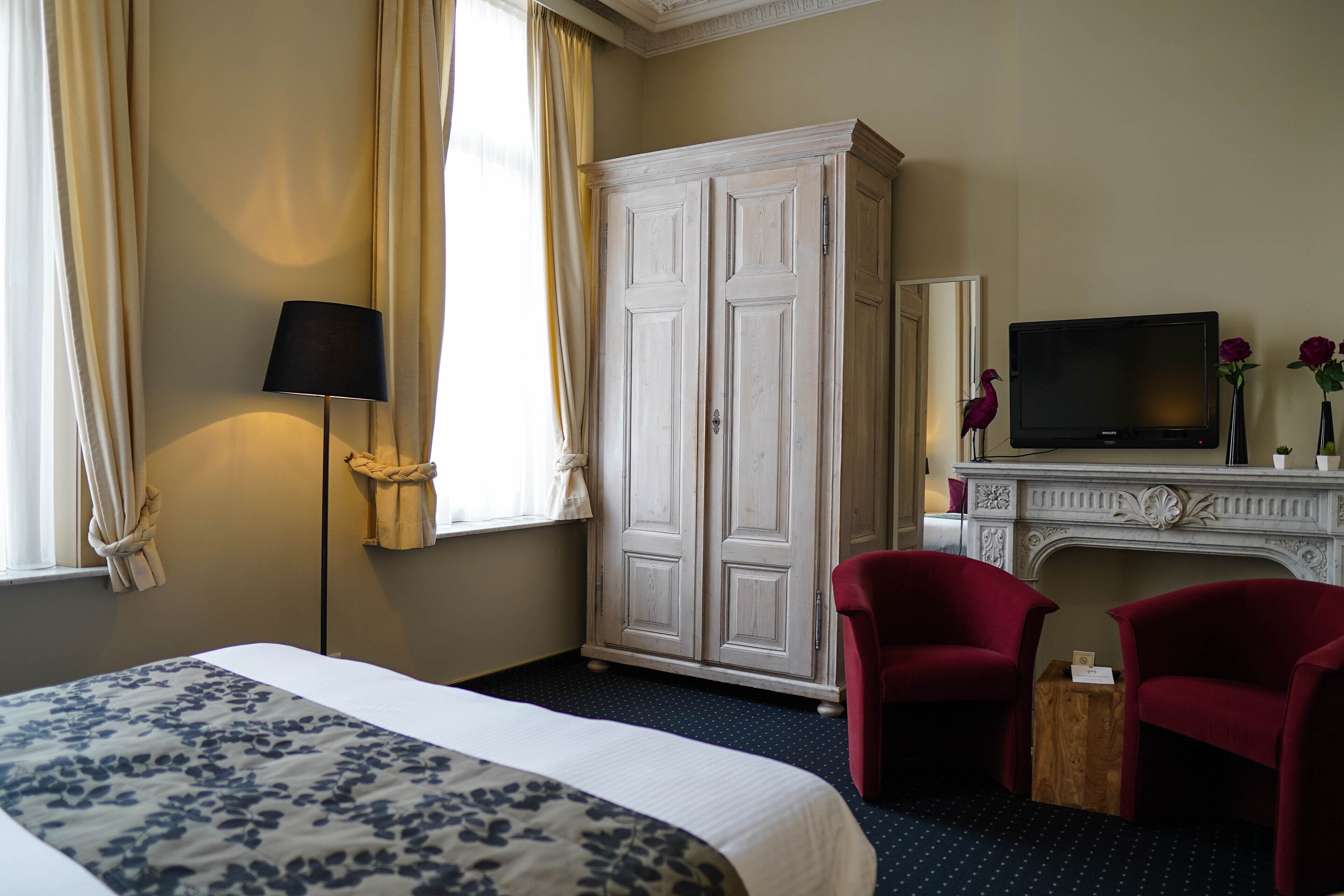 A hotel room with a bed, a television, a wardrobe, two red chairs, and a fireplace.