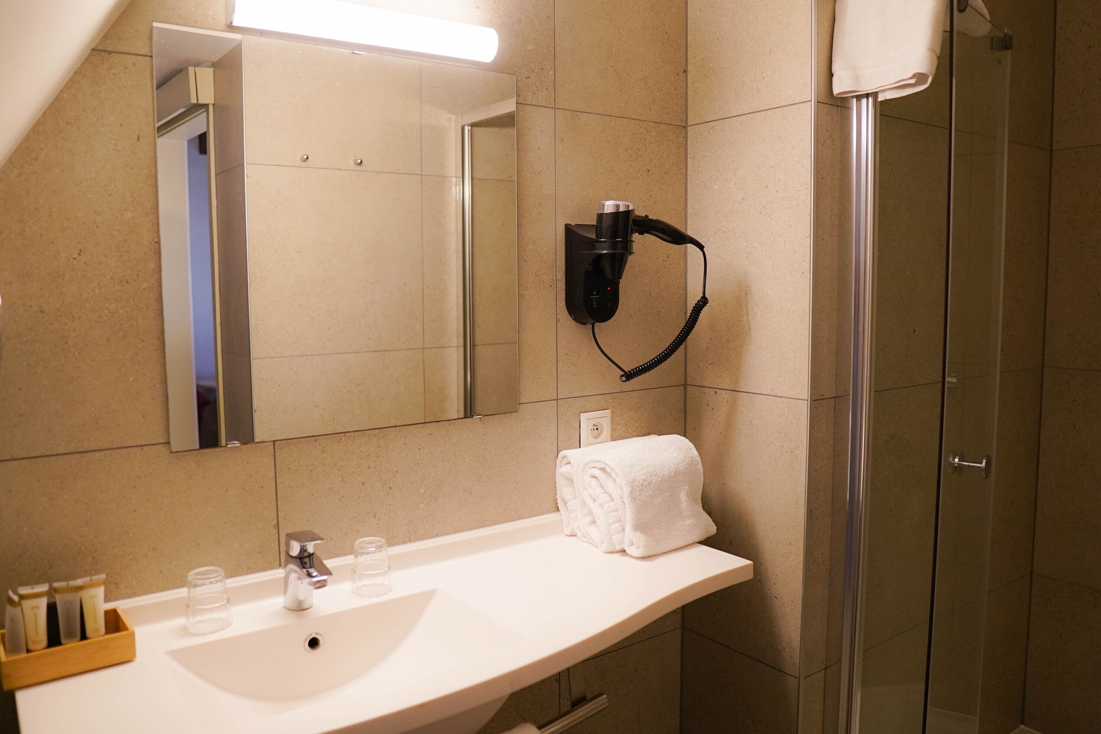 Ein Badezimmer mit einem weißen Waschbecken, einem Spiegel, einem an der Wand montierten Fön und einer Duschkabine aus Glas.