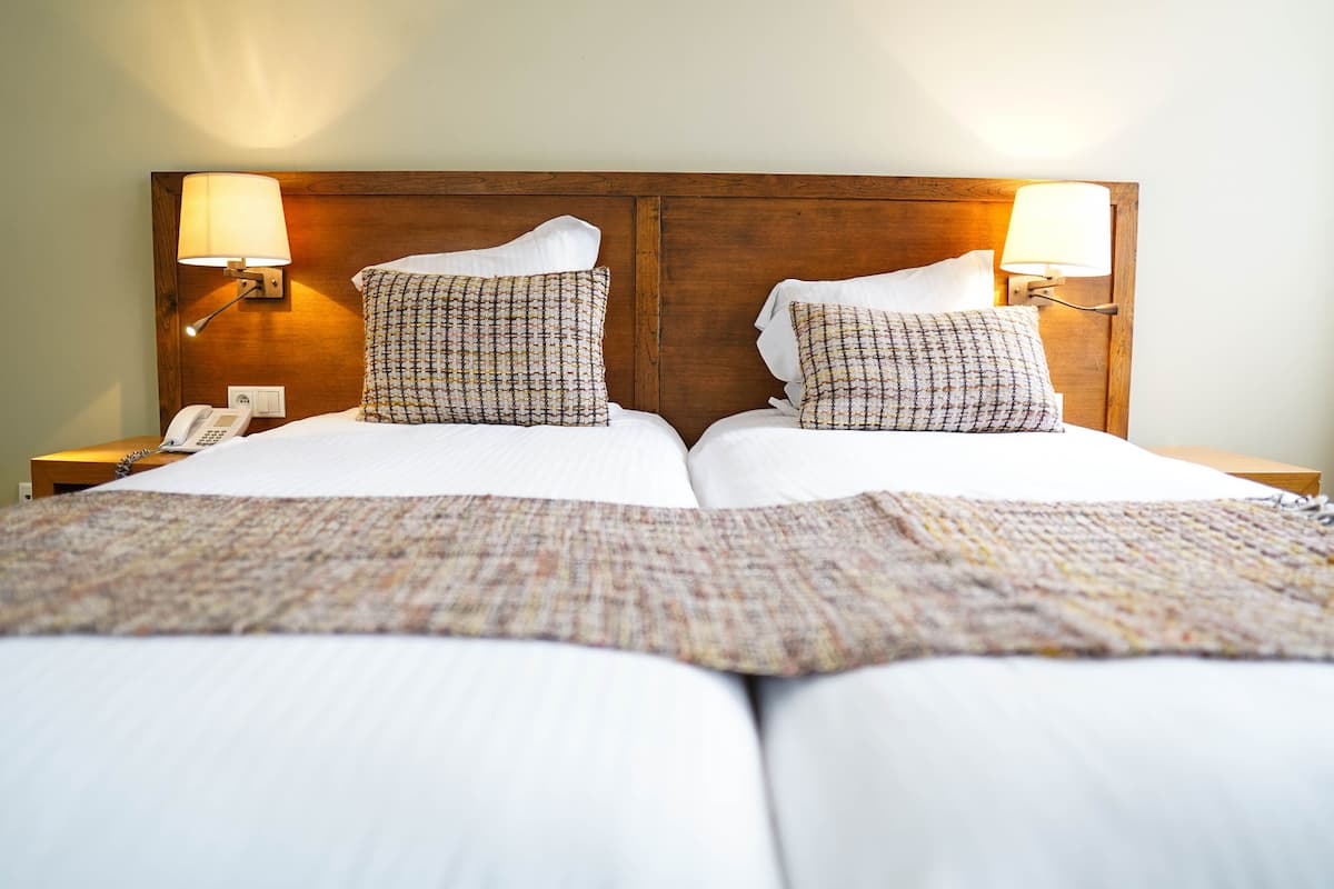 Una cama doble con cabecera de madera, dos almohadas estampadas, una colcha blanca y lámparas de noche.