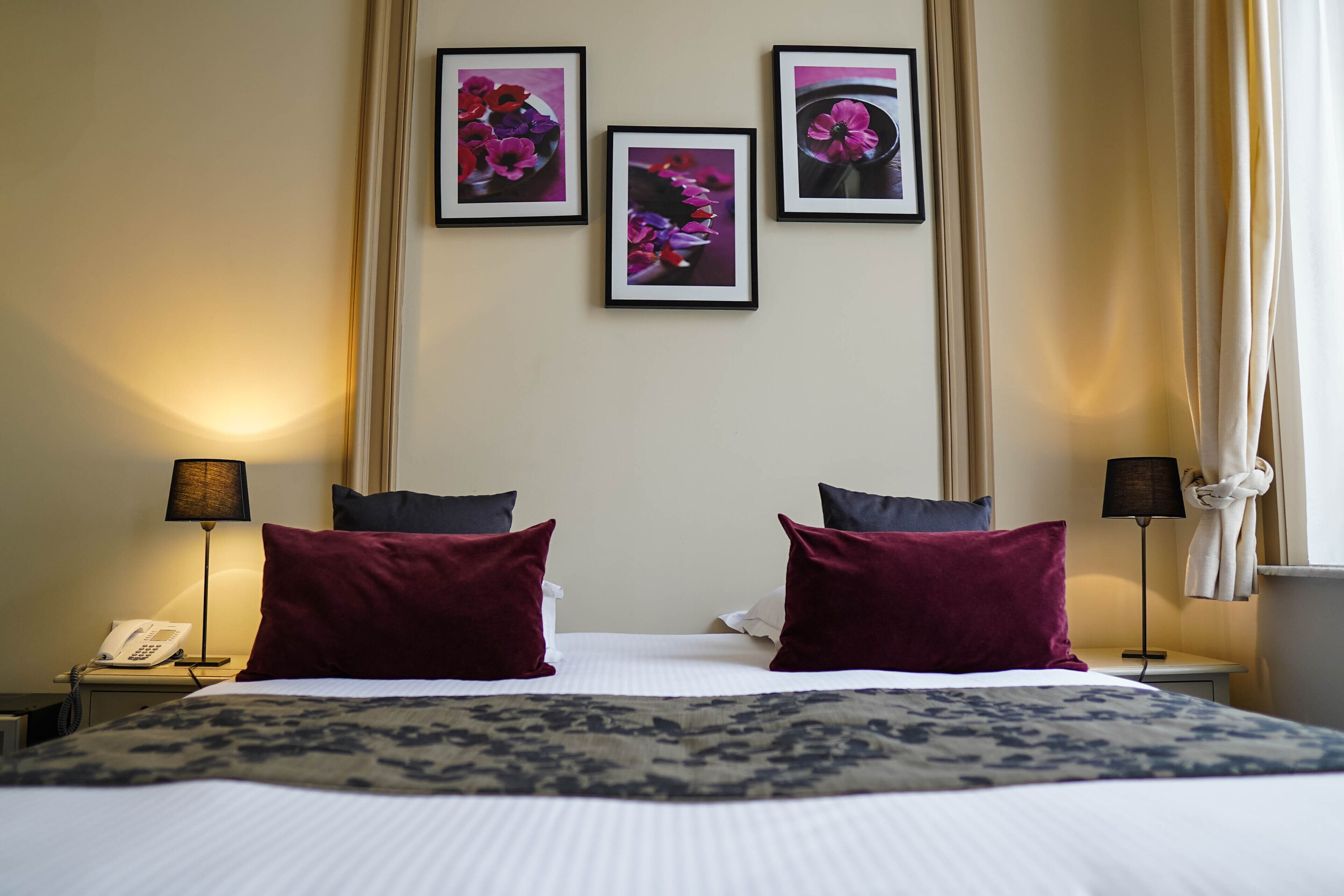 A hotel room with a large bed, two red pillows, a patterned throw, a bedside table with a lamp, a phone, and three framed pictures on the wall.