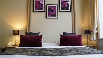 A hotel room with a large bed, two red pillows, a patterned throw, a bedside table with a lamp, a phone, and three framed pictures on the wall.