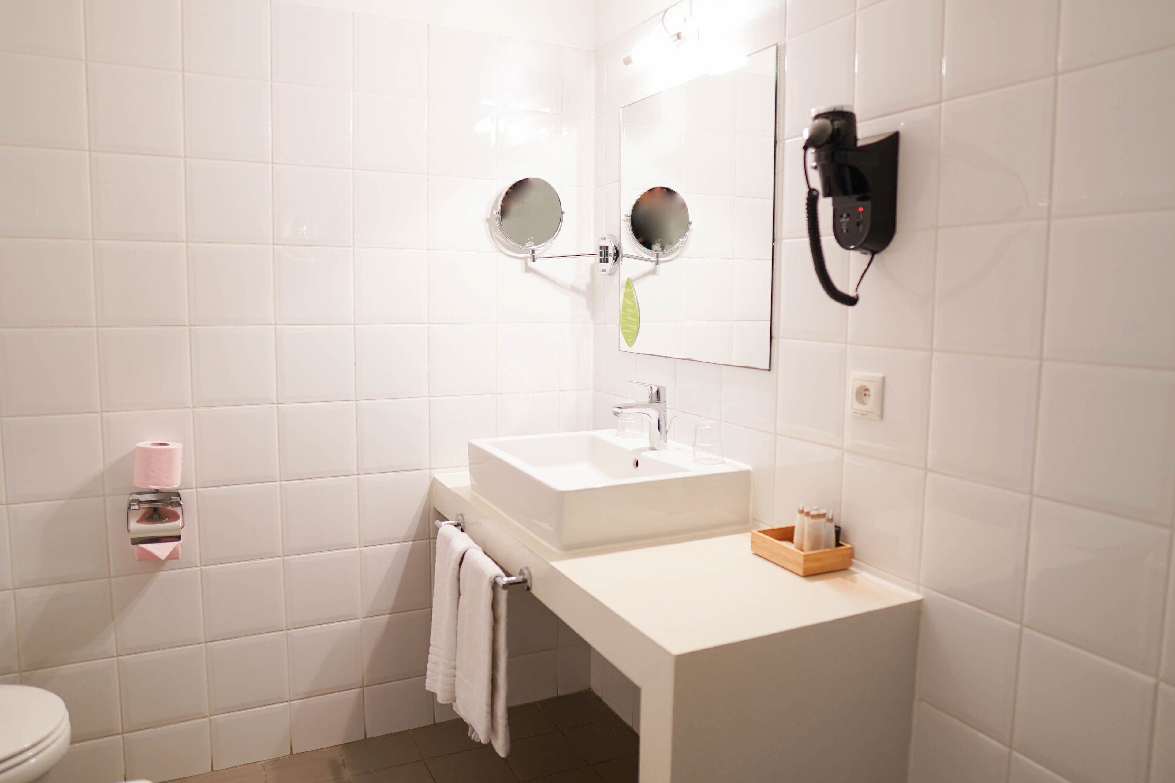 Une salle de bain avec des carreaux blancs, un lavabo, un miroir, un sèche-cheveux et des toilettes.