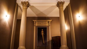 Un couloir avec deux grandes colonnes, un portrait encadré accroché au mur et une porte au fond.
