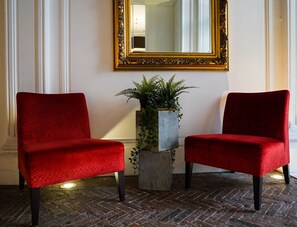 Deux fauteuils rouges à revêtement en tissu, avec des pieds noirs, une plante en pot et un miroir à cadre doré, le tout posé sur un sol en briques.