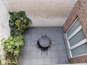 Un petit espace extérieur aménagé en patio, avec une table ronde et des chaises, entouré d’un mur de briques et d’un mur blanc à la texture particulière.