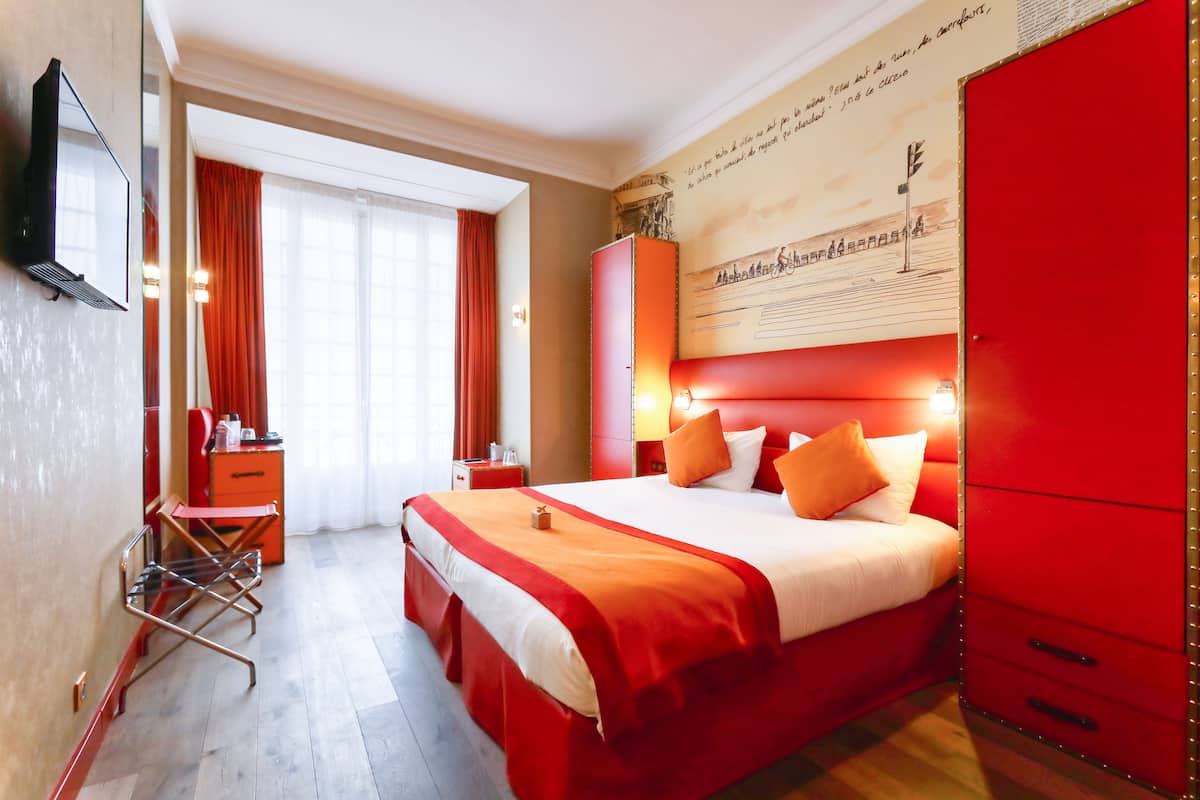 Hotellihuone, jossa on suuri sänky, punainen vaatekaappi, työpöytä tuolilla sekä ikkuna, jossa on verhot.