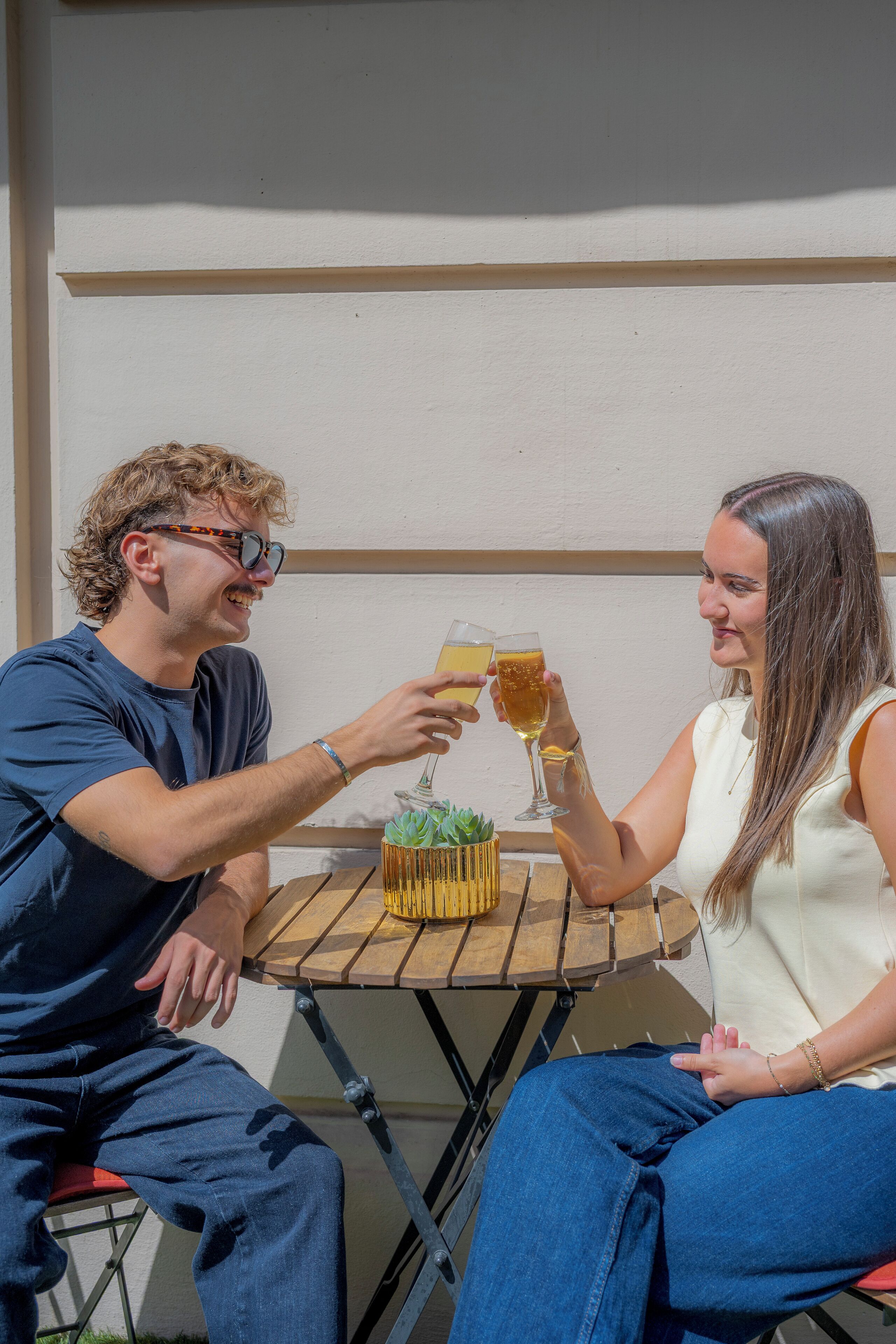 Two people toasting with drinks at a wooden table with a small plant.