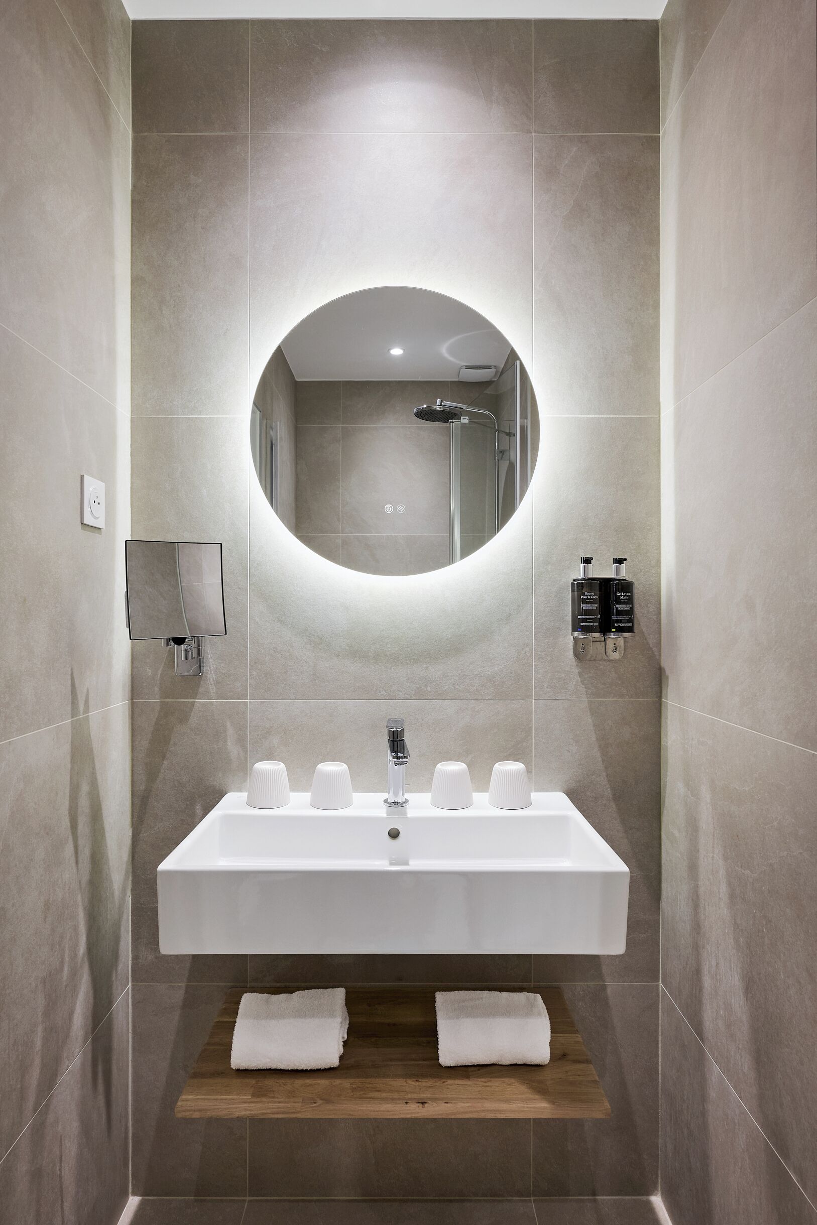 A modern bathroom with a rectangular sink, a round mirror, and two wall-mounted soap dispensers.