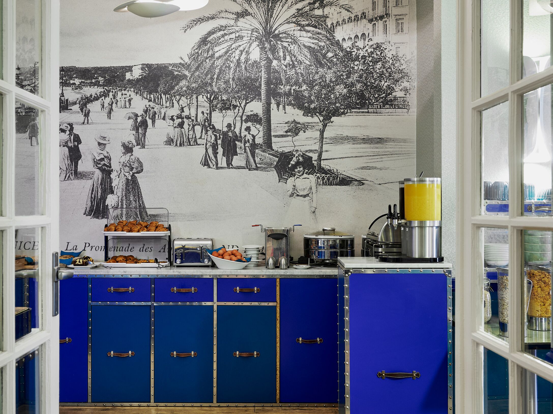 Ein Café im Vintage-Stil mit einer Wandmalerei einer historischen Straßenszene, einer Theke mit Gebäck und blauen Schränken mit Metallgriffen.