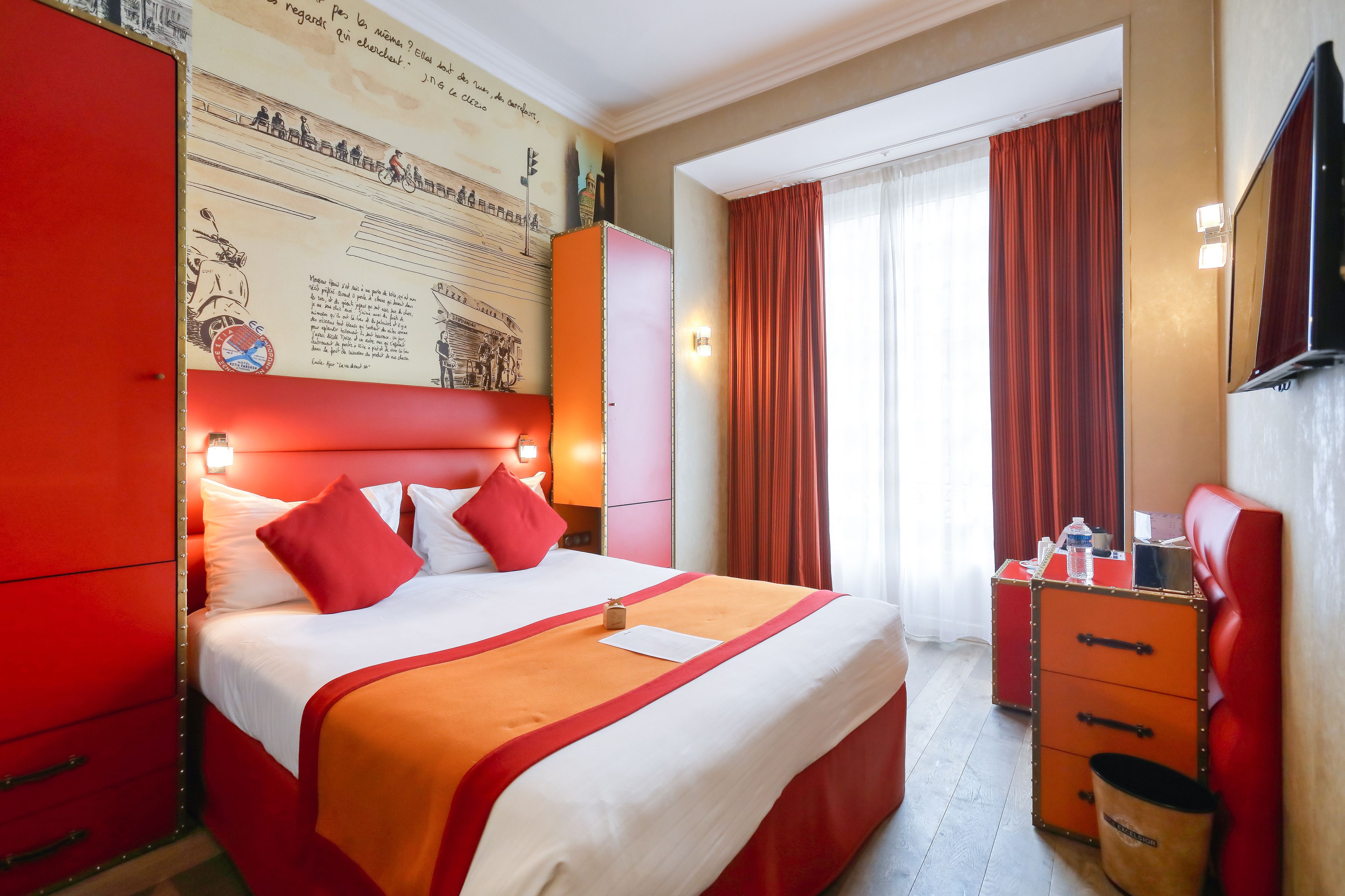A hotel room with a large bed, red and white decor, a desk with drawers, and a window with curtains.