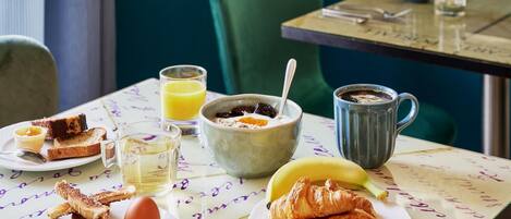 Una mesa de desayuno con tostadas, croissants, un plátano, un tazón de cereal, un vaso de jugo de naranja, una taza de café y una jarra de té.