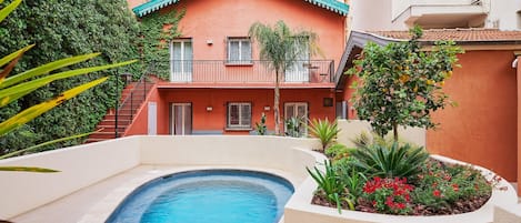 Una piscina rodeada por un jardín con diversas plantas y flores, una escalera que lleva a un balcón, y un edificio con fachada roja y techo verde.