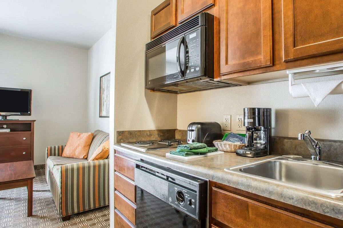 Cocina compacta con microondas, tostadora, cafetera y lavavajillas. Hay un sofá con una manta a rayas y un televisor en la sala contigua.