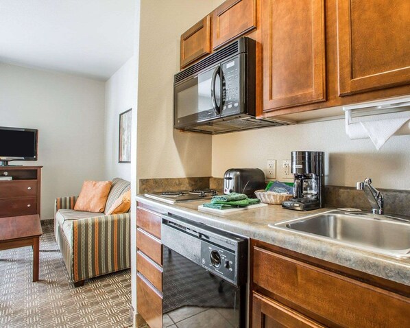 A compact kitchen with a microwave, toaster, coffee maker, and dishwasher. There is a sofa with a striped blanket and a television in the adjacent living area.