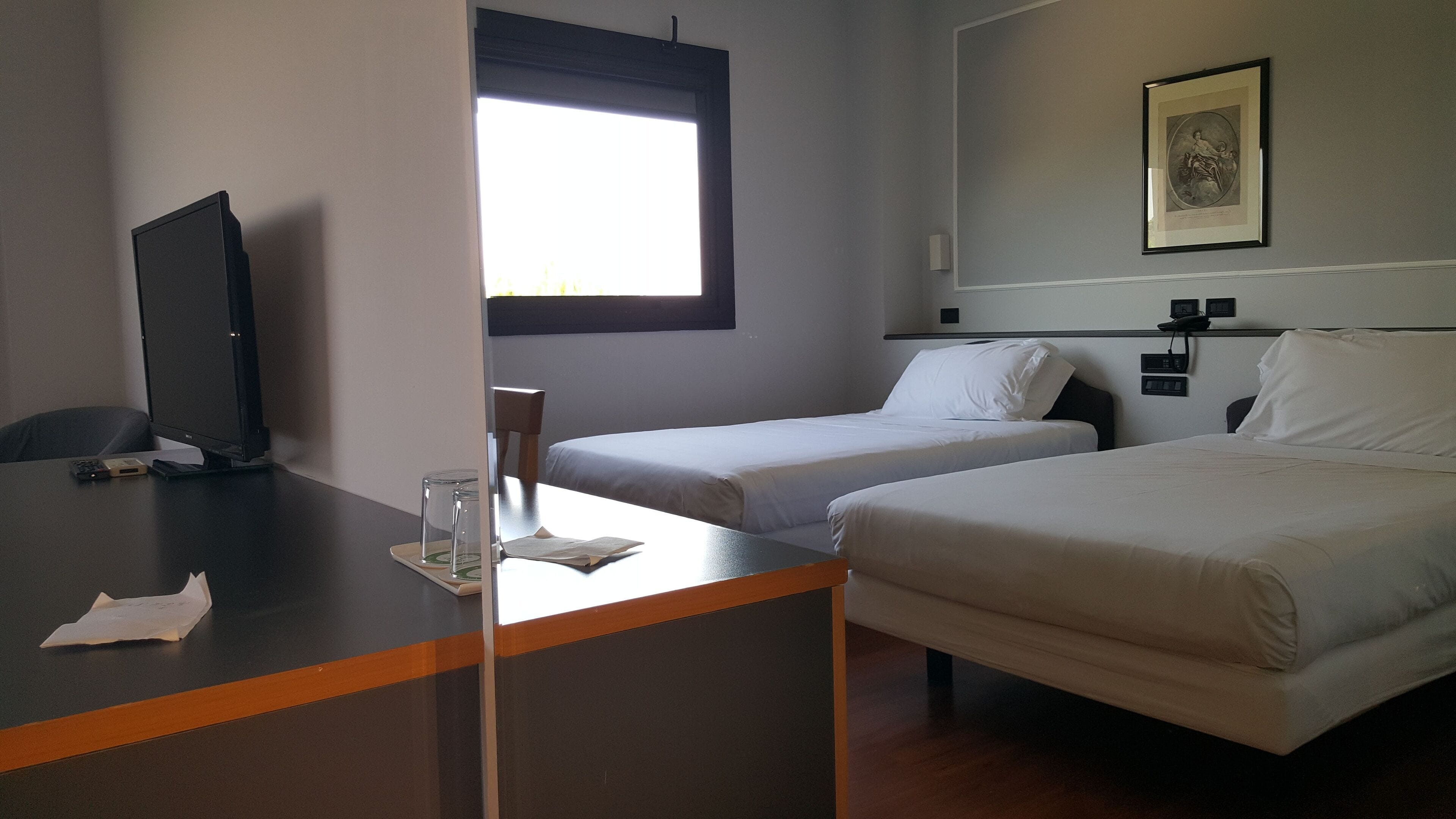 Ein Hotelzimmer mit Schreibtisch, zwei Einzelbetten, einem Fenster, einem gerahmten Bild an der Wand und einem Fernseher auf dem Schreibtisch.