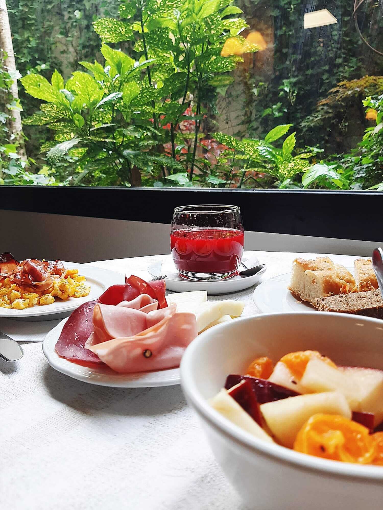 A breakfast setting with plates of food, a glass of red beverage, and a view of greenery through a window.