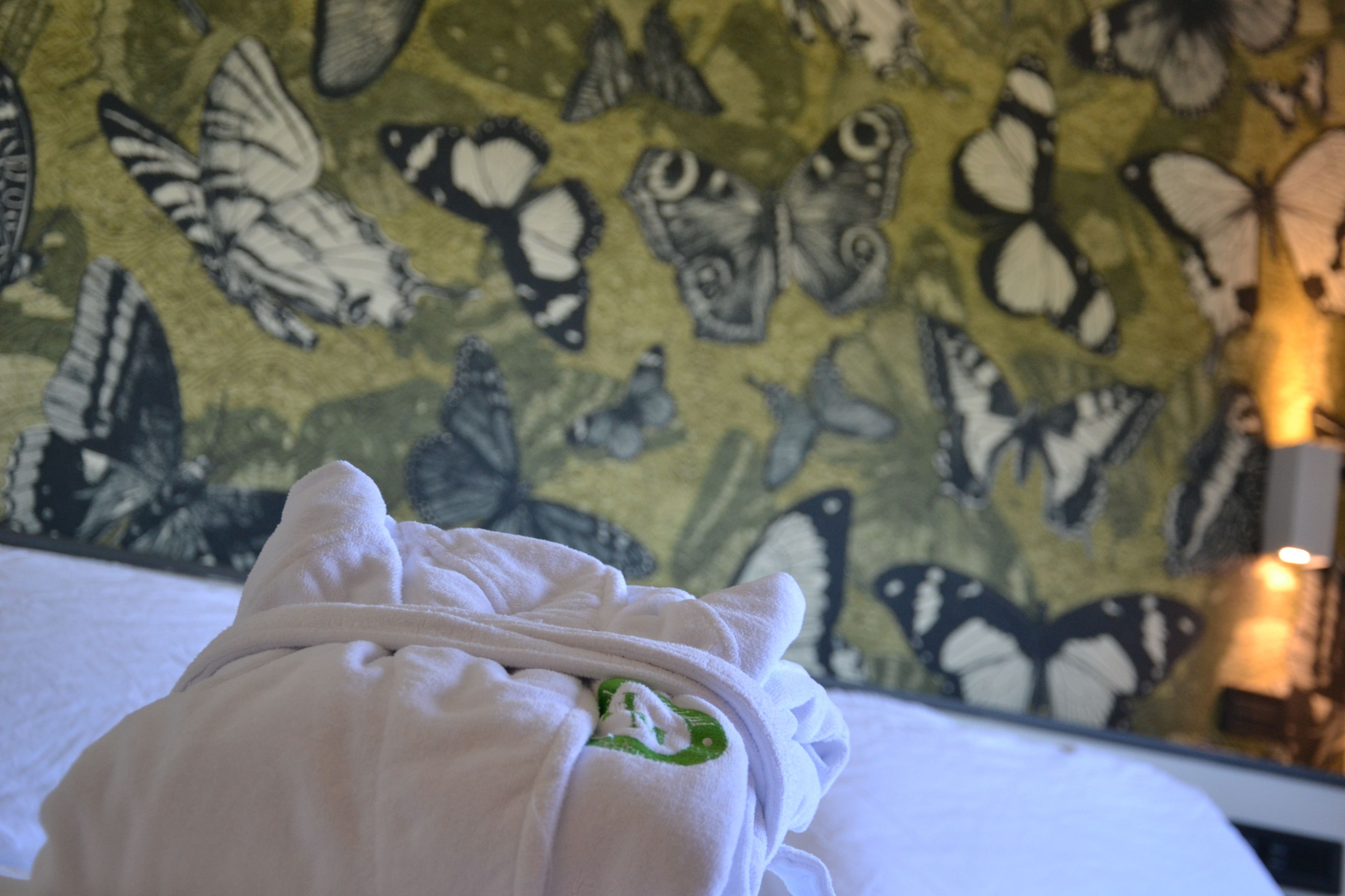 Una cama con una manta blanca y un emblema verde, frente a una pared con estampado de mariposas.