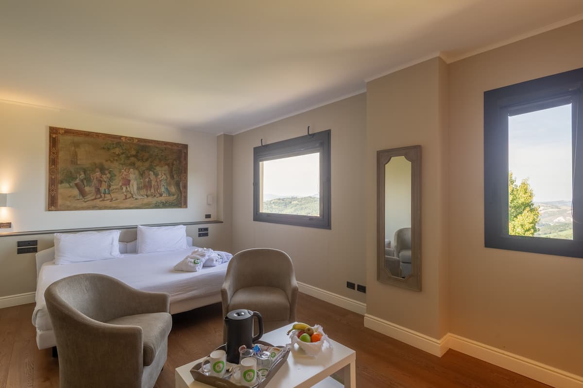 A hotel room with a bed, two armchairs, a coffee table with a tray of mugs and a fruit bowl, a painting on the wall, and two windows with views.
