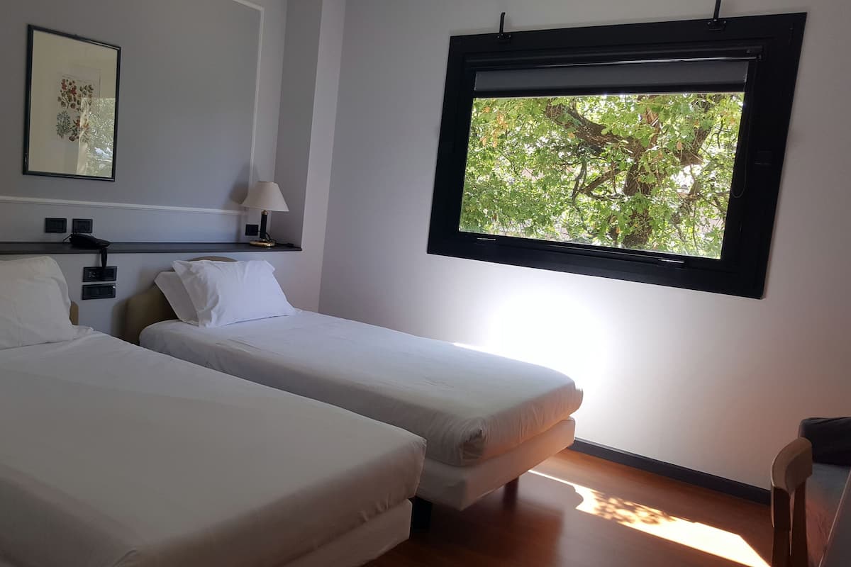 A hotel room with two single beds, a bedside table with a lamp, a framed picture on the wall, and a window with a view of trees.