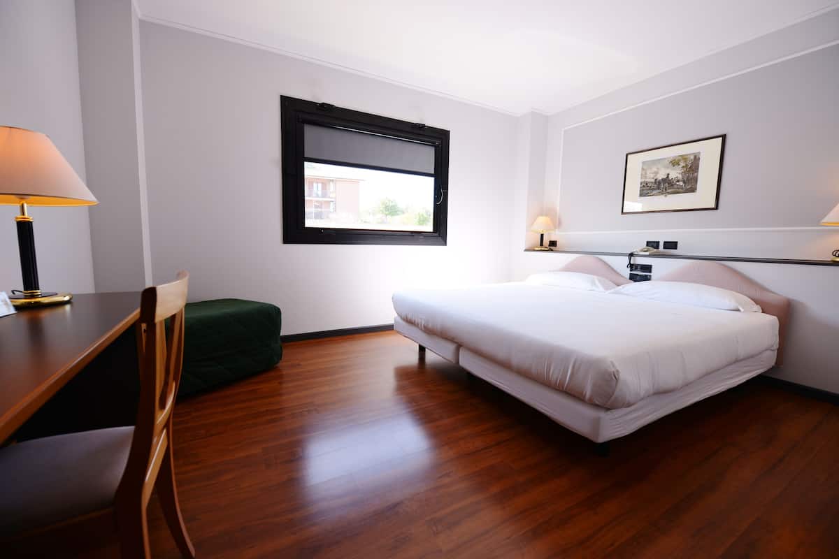 A hotel room with a bed, a desk, a chair, a window, and a framed picture on the wall.