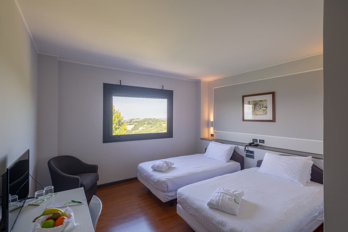 A hotel room with two beds, a desk with a chair, a window with a view, and a framed picture on the wall.