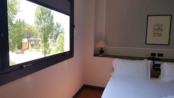 Quarto de hotel com cama, mesa de cabeceira, luminária e janela com vista para árvores e um carro do lado de fora.