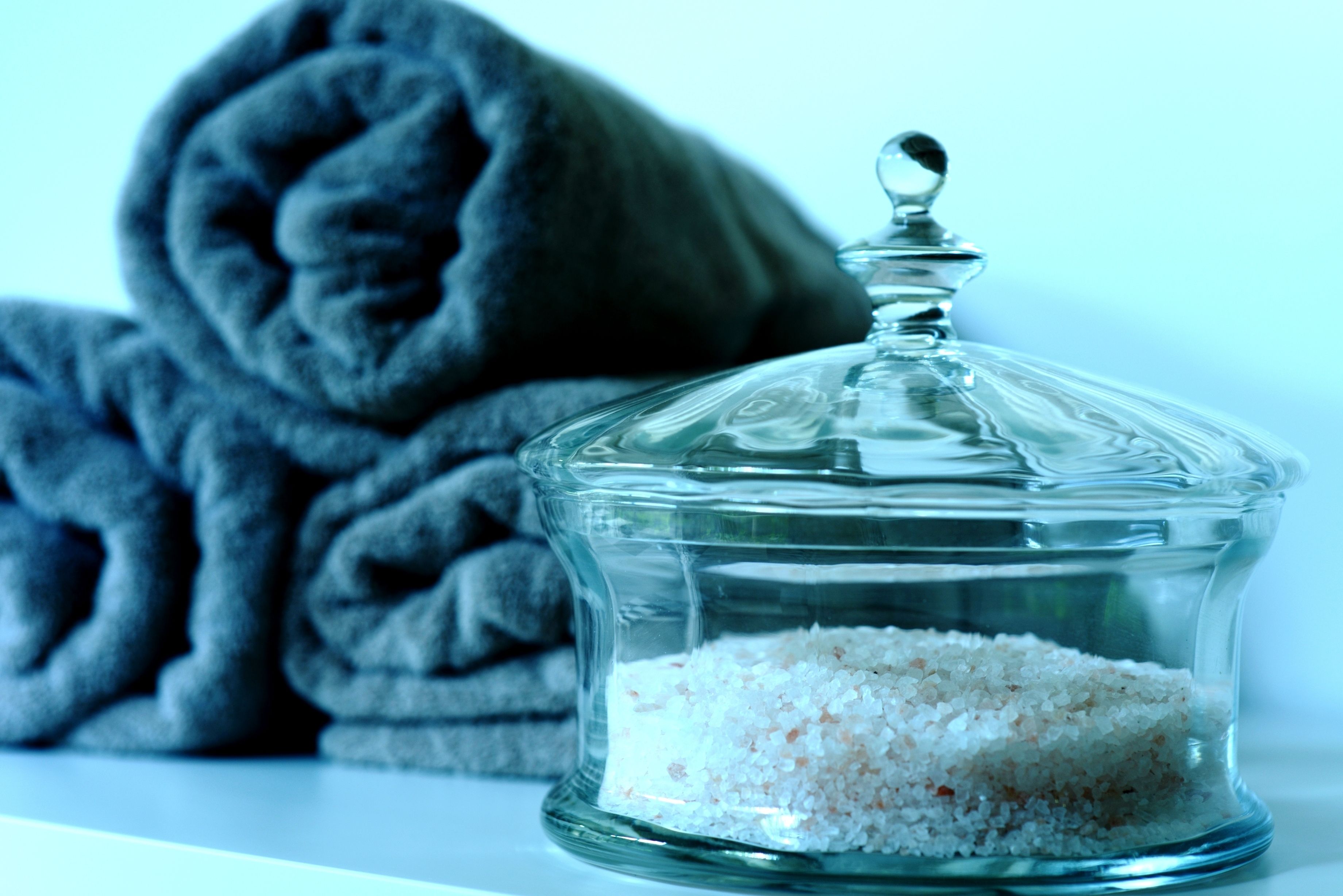 A glass container with white bath salts and a rolled towel in the background.