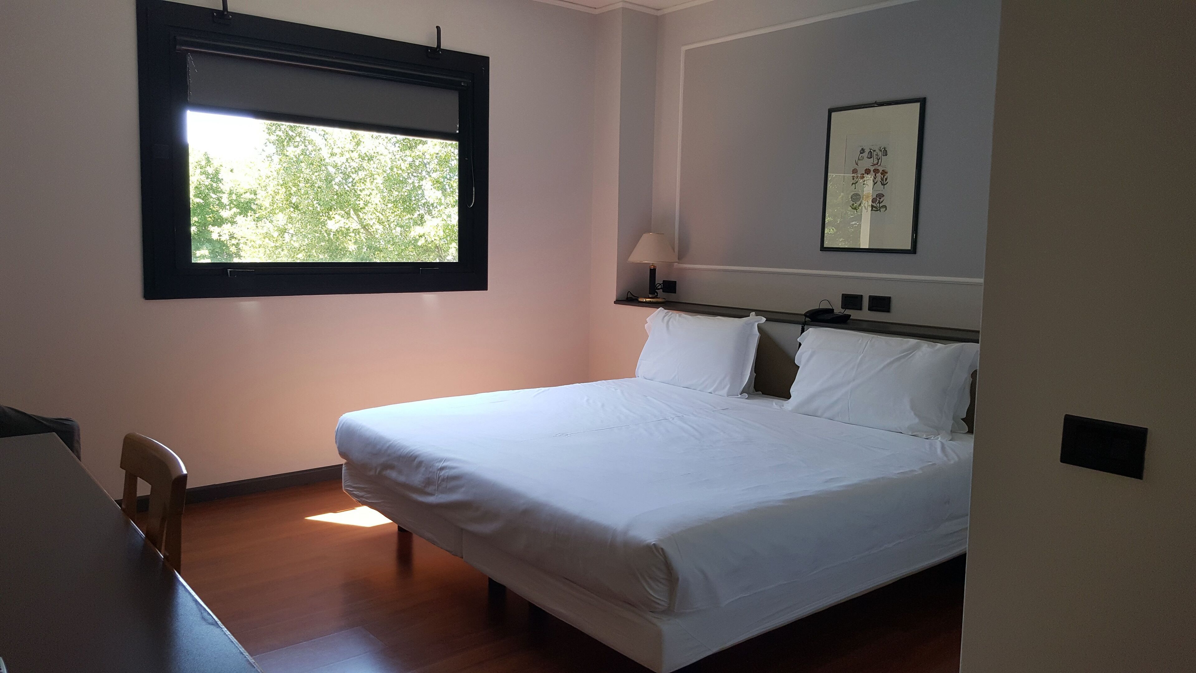 A hotel room with a bed, a desk, a chair, a window, and a wall-mounted lamp.