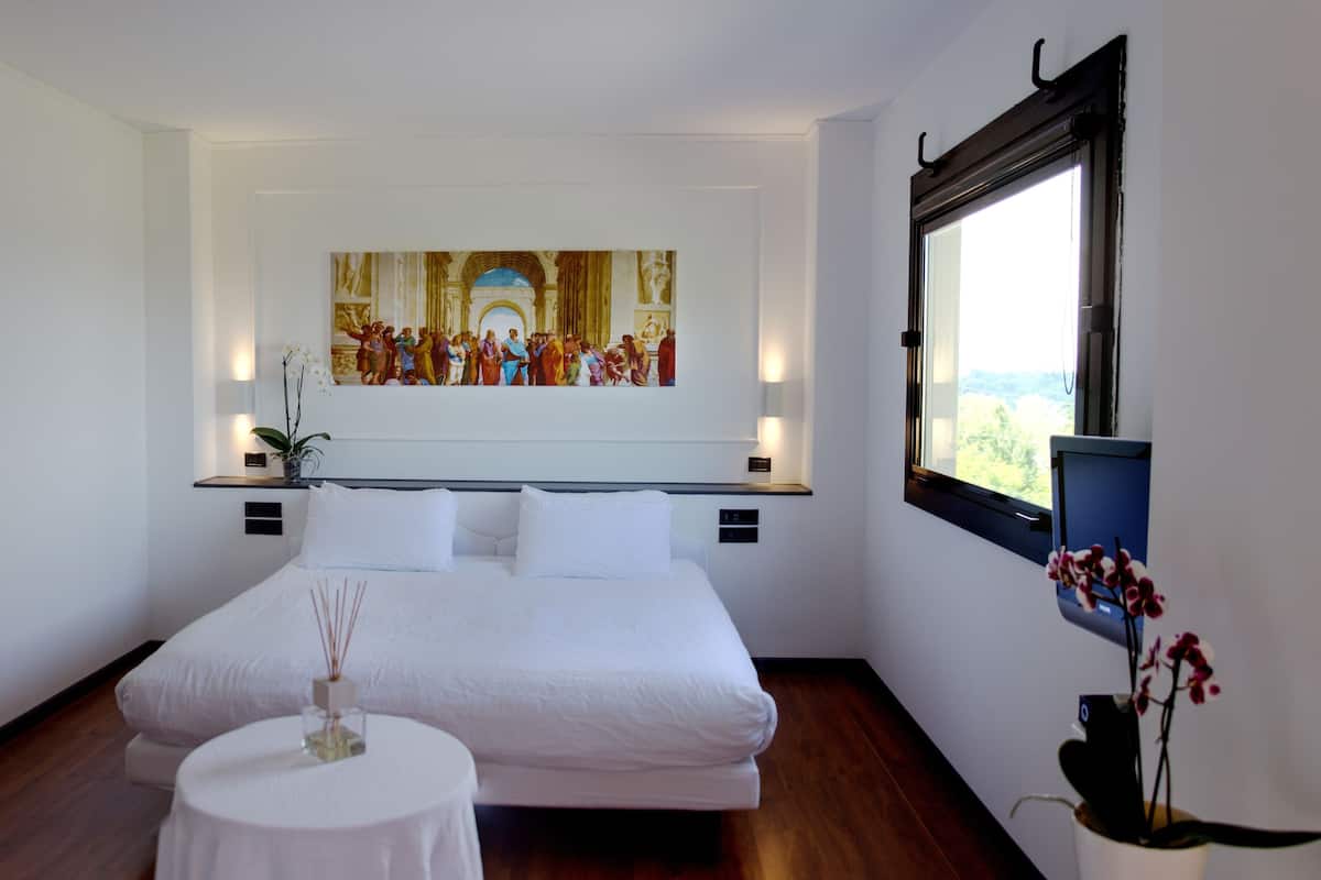 A bedroom with a large bed, a small round table, a potted plant, and a painting on the wall.