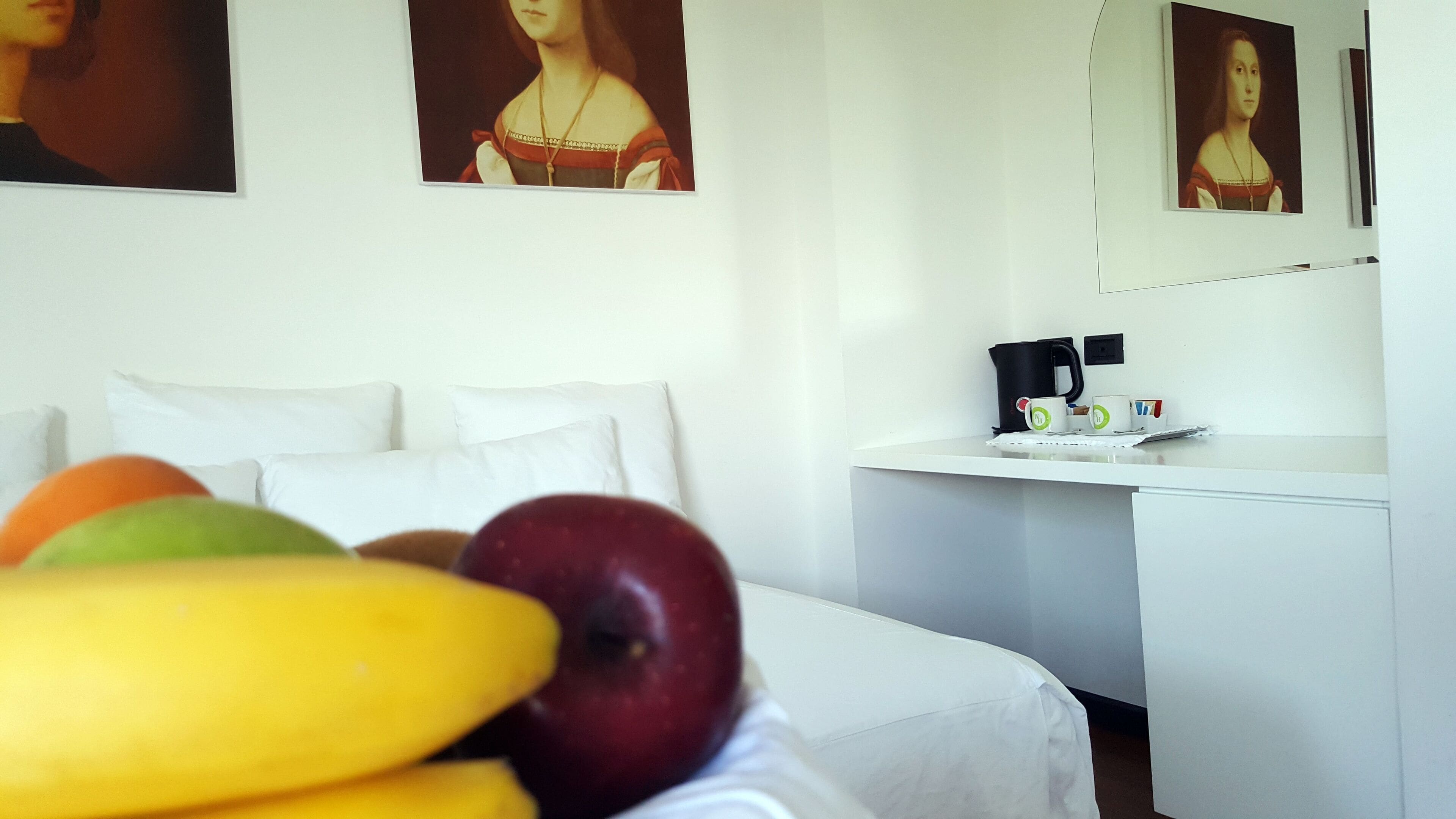 Hotellihuone, jossa on sänky, yöpöytä, jossa on vedenkeitin ja kuppeja, sekä hedelmiä sängyllä.