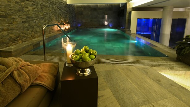 Zona de spa con pared de piedra, piscina, toalla, un cuenco de manzanas y una vela.