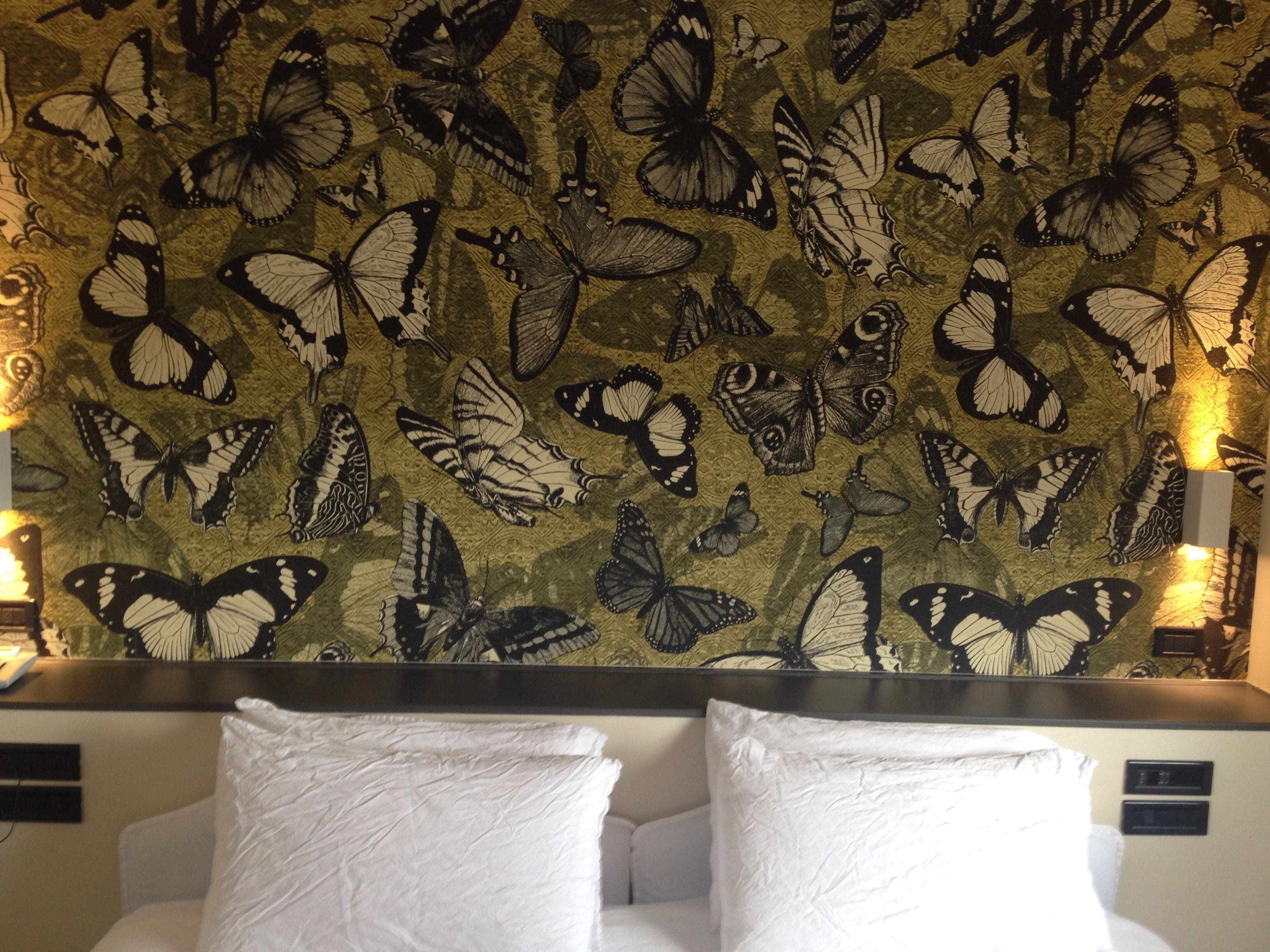 A hotel room with a wall decorated with a butterfly pattern. There are two white pillows on the bed.