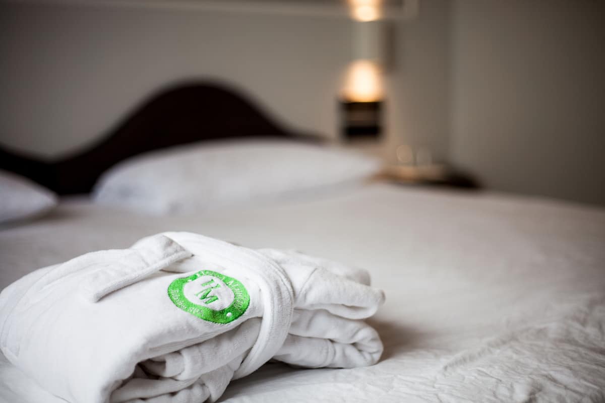 A neatly made bed with a white blanket and a towel with a green logo.