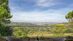 Un balcón con sillas de mimbre y una mesa, con vistas a un paisaje pintoresco con colinas y árboles.