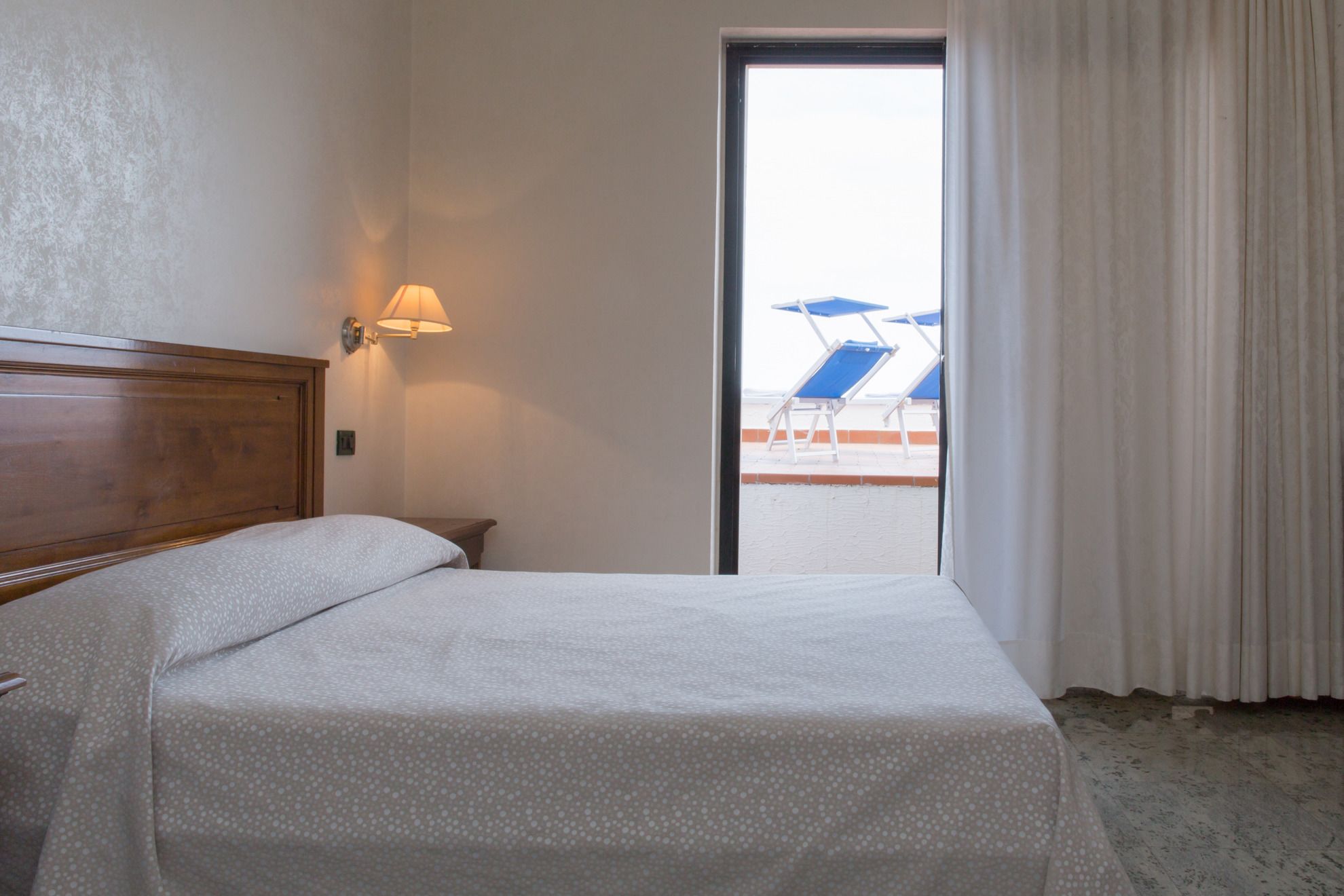 A hotel room with a bed, a nightstand, a lamp, and a view of a pool area with blue chairs and umbrellas.