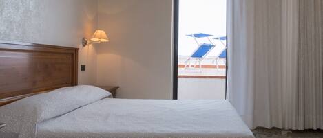 Habitación de hotel con cama, mesita de noche, lámpara y vista a un área de piscina con sillas y sombrillas azules.