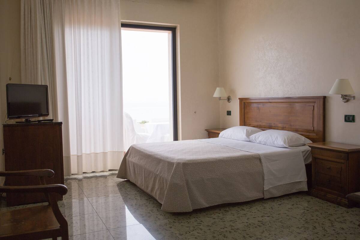 Habitación de hotel con cama, televisión sobre un mueble de madera, una silla, mesita de noche con lámpara y ventana con cortinas.