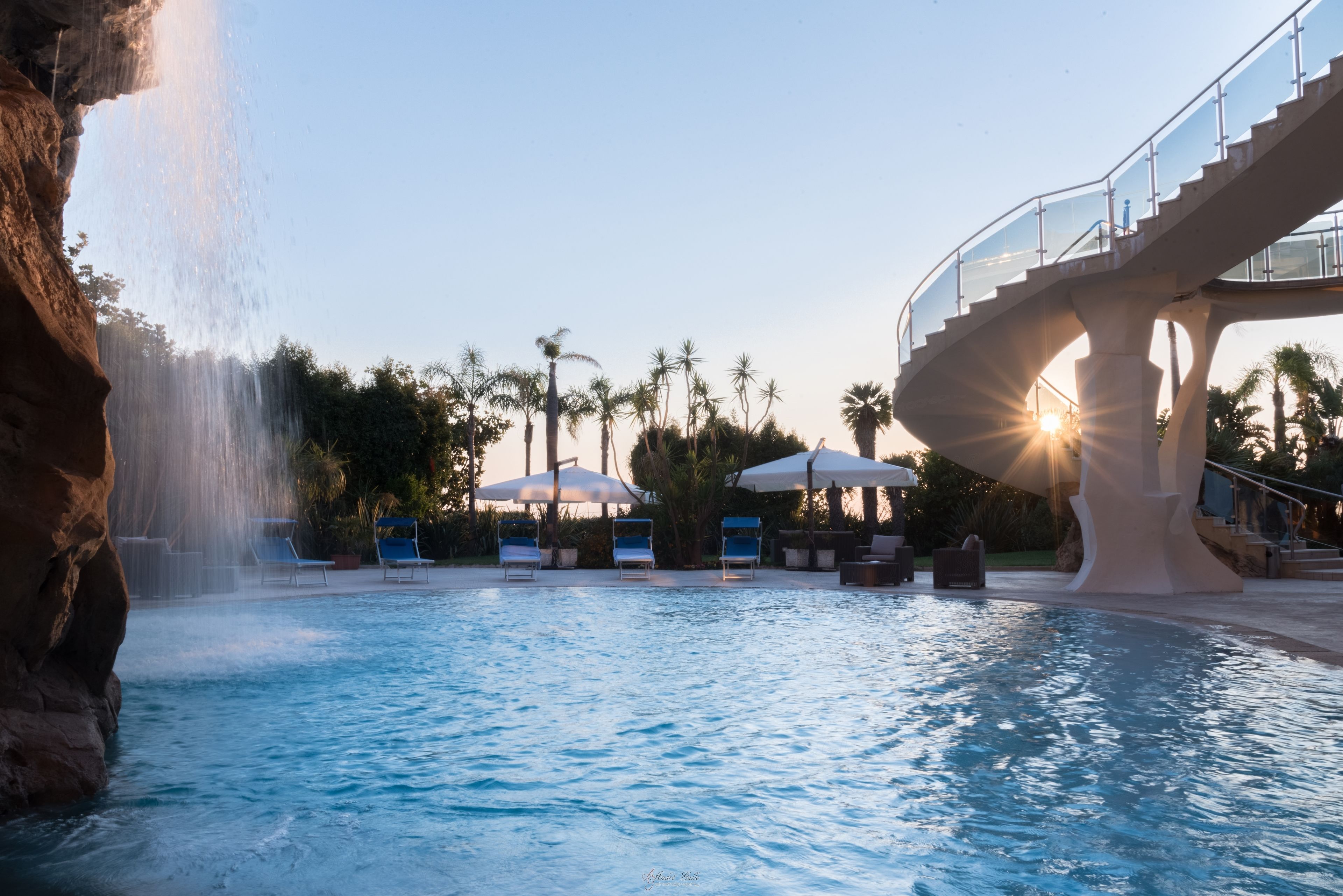 A pool with a waterfall feature, lounge chairs, umbrellas, and a glass bridge.