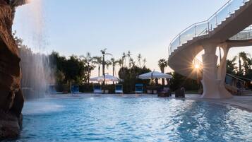 A pool with a waterfall feature, lounge chairs, umbrellas, and a glass bridge.
